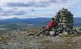 A person sitting at a summit cairn taking in a far view over mountains, valleys and lakes.