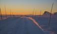 Brøytet vei i solnedgang. Brøytestikker og en topp horisonten stikker opp fra snøen.
