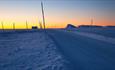 Eine Straße zieht sich durch Schneelandschaft zu Beginn der Blauen Stunde, während das Restlicht der Sonne den Horizont gelb-orange färbt.