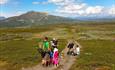 To familier på en bred sti på snaufjellet om sommeren med et fjell i bakgrunnen.