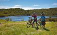 Two cyclists walk with their bikes over green grass in front of a blue lake.