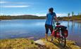 Female cyclist with a blue cycling shirt stands next to her bike looking out on a blue lake.