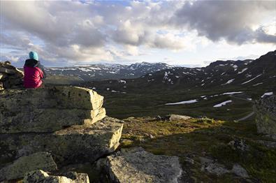 En person sitter på en stor stein og ser ut over fjellandskap i motlys