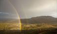 Dobbel regnbue over en åpen sadel med vann, lyng og høyvoksende granskogspletter mellom to fjelltopper.