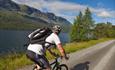 A cyclist cycles on a gravel road alongside a lake and looks to the mountains on the opposite shore.