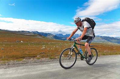 Syklist på vei opp bakkene på Slettefjellveien med Jotunheimens tinder i bakgrunnen.