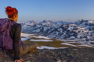 Eine Frau sitzt im Vordergrund und genießt die Aussicht zum Hochgebirge mit vielen Schneefeldern im lila Morgenlicht.