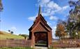 A stave church beyond  a wooden fence with a portal with ornaments. Blue skies.