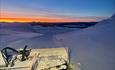 A grooming machine for cross-country skiing tracks in teh mountains before sunrise with an orange band in the sky over the horizon.
