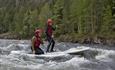 Two stand-up boarders, one on her knees and one standing upright, navigating a rapid in the river