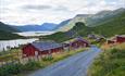 Ein Almweg führt durch die idyllische Almsiedlung Strø mit ihren roten Hütten. Im HIntergrund liegen Seen und Berge.