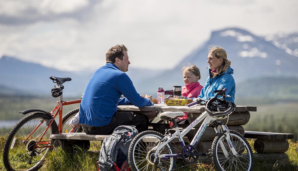 Eine Familie auf Fahrradtour macht eine Rast an einem Picknicktisch. Berge im Hintergrund.