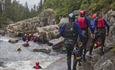 A group of persons with wetsuits and helmets walks and swims downstream through rapids in a river.