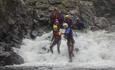 A group of persons with wetsuits and helmets walks downstream in rapids in a river.
