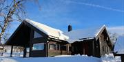 Hytte med snø rundt og på taket, i sol og fint, klart vintervær.