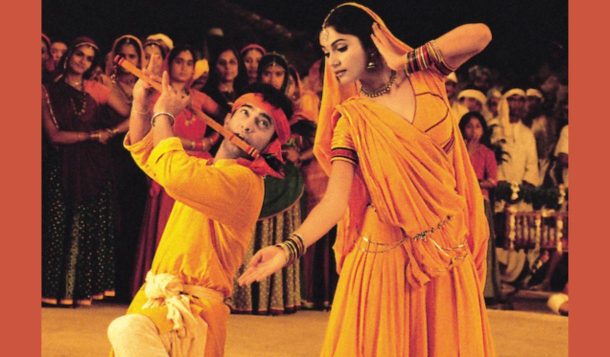 A picture of a man and woman in traditional Indian dress. She is dancing, and he is playing the flute