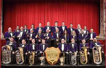 A photograph of Brighouse and Rastrick Brass Band, on a stage, with their instruments, plus a shield and two trophies.