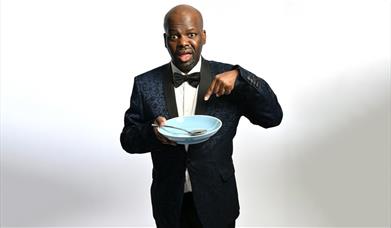 A picture of comedian Daliso Chaponda, holding, and pointing at, an empty bowl and spoon