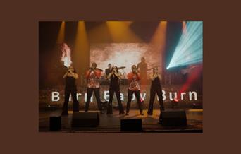 A picture of five people singing on stage, with the words "Burn Baby Burn" visible on a backdrop behind them