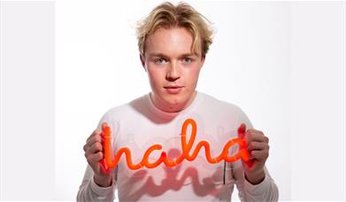 A picture of comedian Finlay Christie holding an orange neon sign which says "haha"