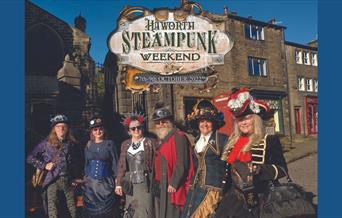 A picture of six people on steampunk costumes, standing together in front of shops on a cobbled street