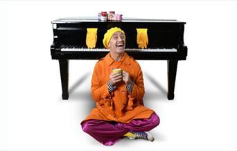A picture of a man wearing an orange mac and a yellow beret, sitting cross-legged, in front of a grand piano