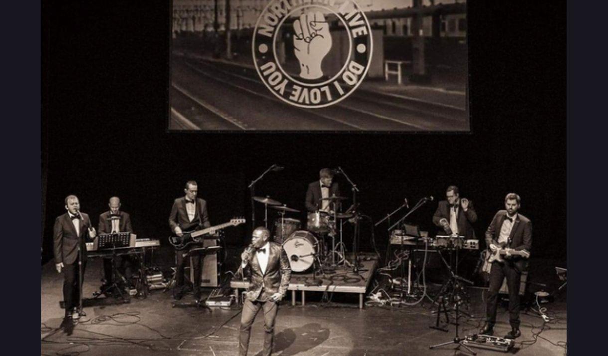 A black and white picture of a seven-piece soul band, performing on stage, in front of a screen with their band logo on it