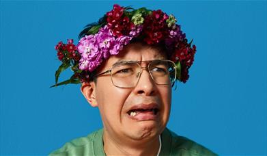 A picture of comedian Phil Wang, looking upset. He is wearing a crown of flowers on his head.