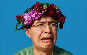 A picture of comedian Phil Wang, looking upset. He is wearing a crown of flowers on his head.