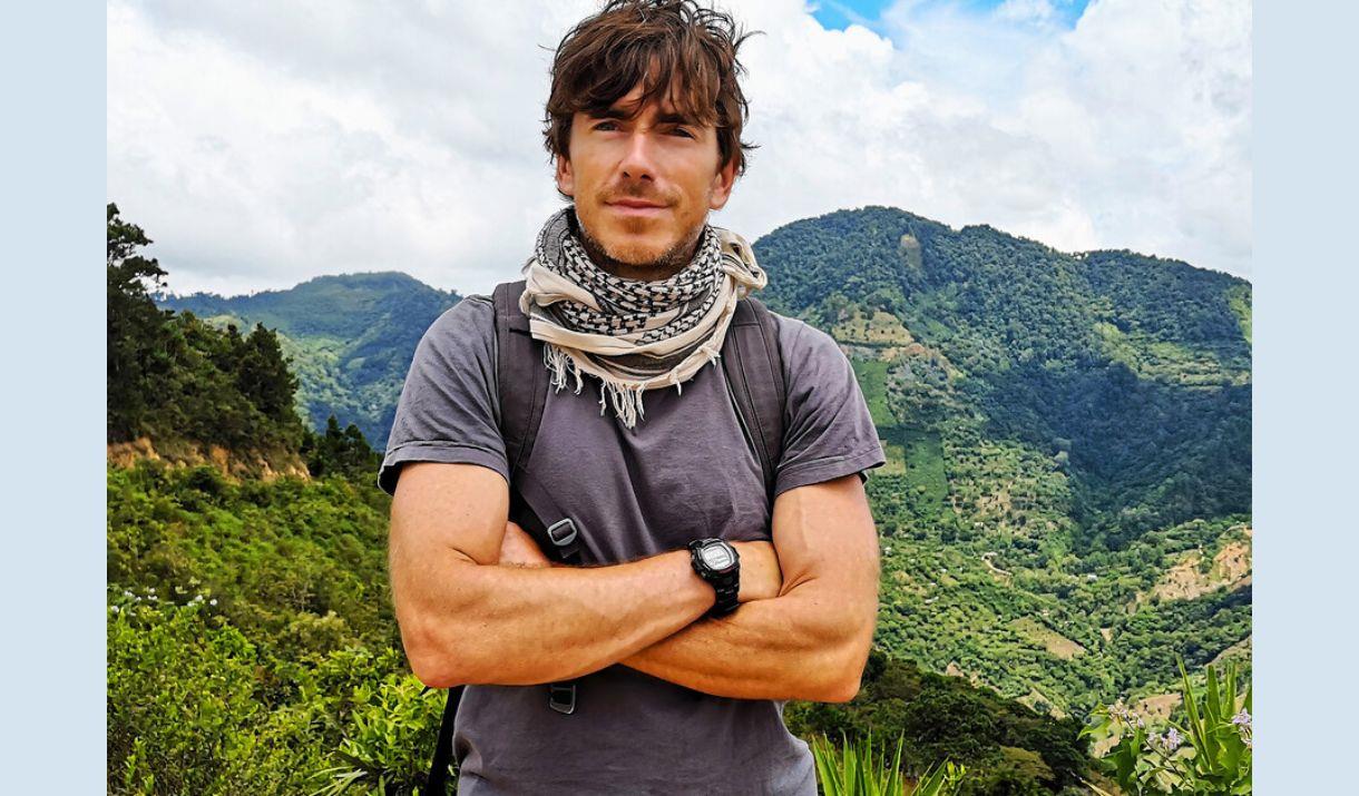 A picture of Simon Reeve, against a backdrop of forested green hills