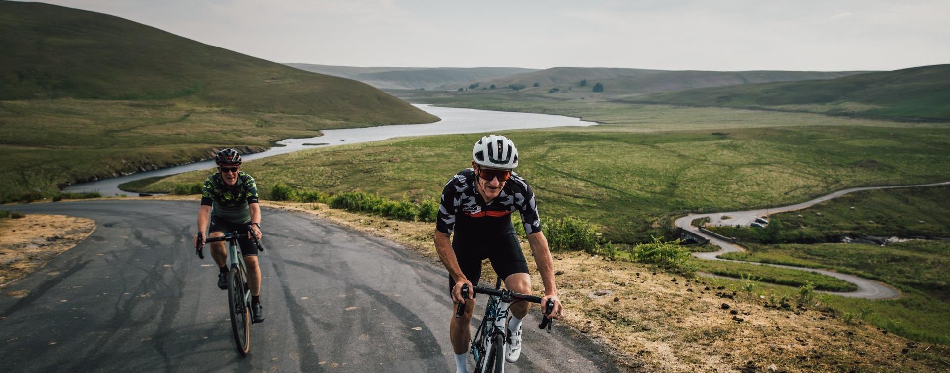 Two cyclists climbing a mountain road above the winding Elan Valley reservoirs