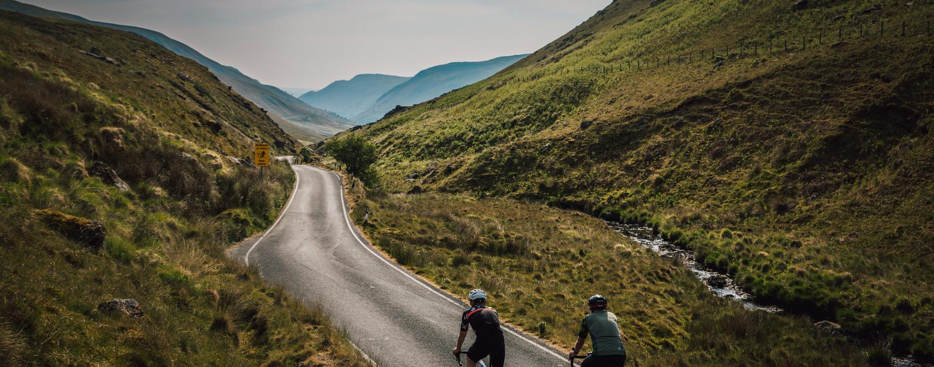 Two road cyclists riding down a narrow road through a steep green valley in the Cambrian Mountains
