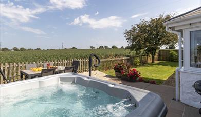 The beautiful, bubbling hot tub is in the foreground with the lawn stretching away. The patio dining table and chairs overlook the countryside