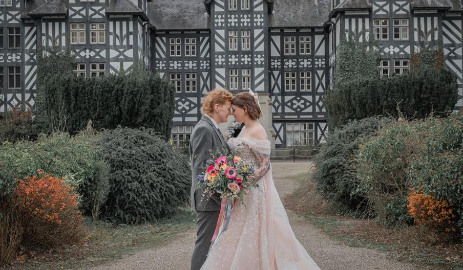 Two women in wedding outfits in front of a black and white stately home