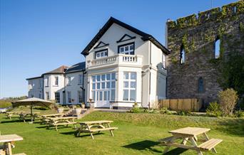 A Stunning White Hotel (The Castle Of Brecon Hotel) & The Medieval Castle Wall. The Sky Is Blue And there Are Light Wooden Benches On The Green Grass,