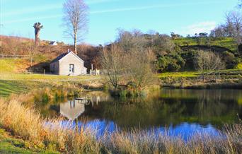 A stone and slate barn sits across from a pond, its reflection clear on water. Trees and grass surround the pond