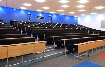 Empty lecture room with rows of seats and desks facing a screen.