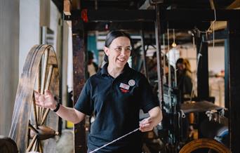 A woman dressed in a black polo shirt is spinning yarn on the Great Wheel in a gallery at the National Wool Museum