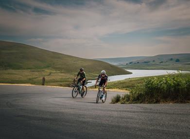 Two cyclists on an open road above a river valley in the hills of Mid Wales