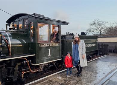 A mother and child beside a steam engine as the driver in a Santa hat waves from the cab