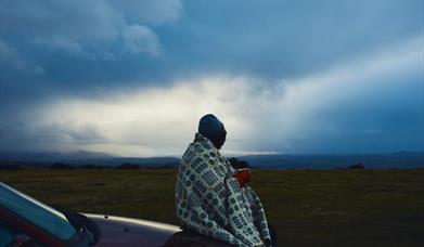 A person wrapped in a Welsh wool blanket holding a warm mug at dusk