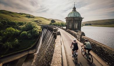 Cyclists riding across the top of a Victorian dam in the Elan Valley with water pouring over the side