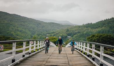 Three cyclists riding towards the camera over the wooden toll bridge at Penmaenpool on the Mawddach Trail