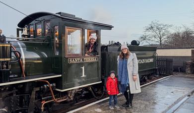 A mother and child beside a steam engine as the driver in a Santa hat waves from the cab