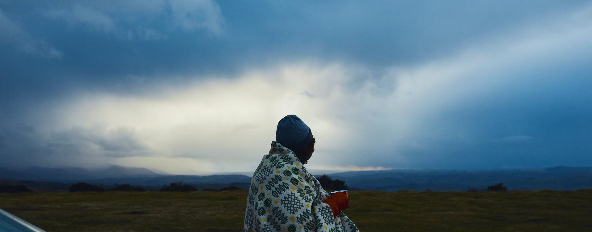 A person wrapped in a Welsh wool blanket holding a mug on Hay Bluff as evening falls