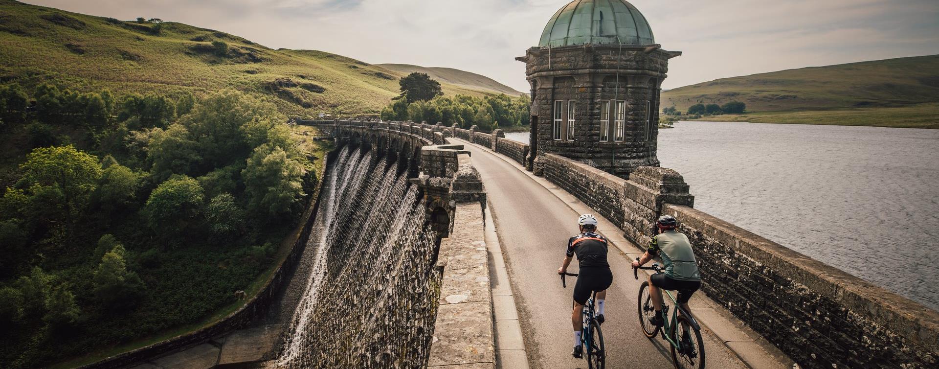 Two road cyclists riding across Craig Goch dam in the Elan Valley on Lôn Cambria