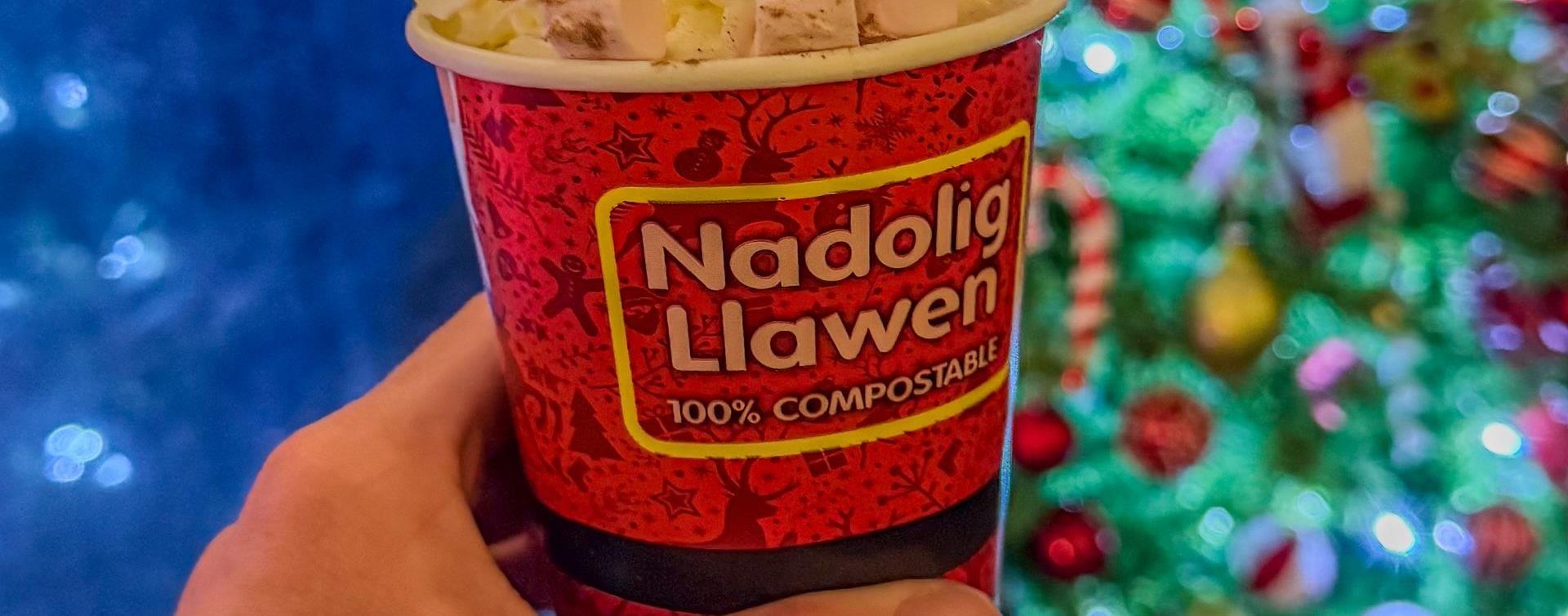 A hot chocolate with marshmallows in a Nadolig Llawen cup in front of a decorated Christmas tree