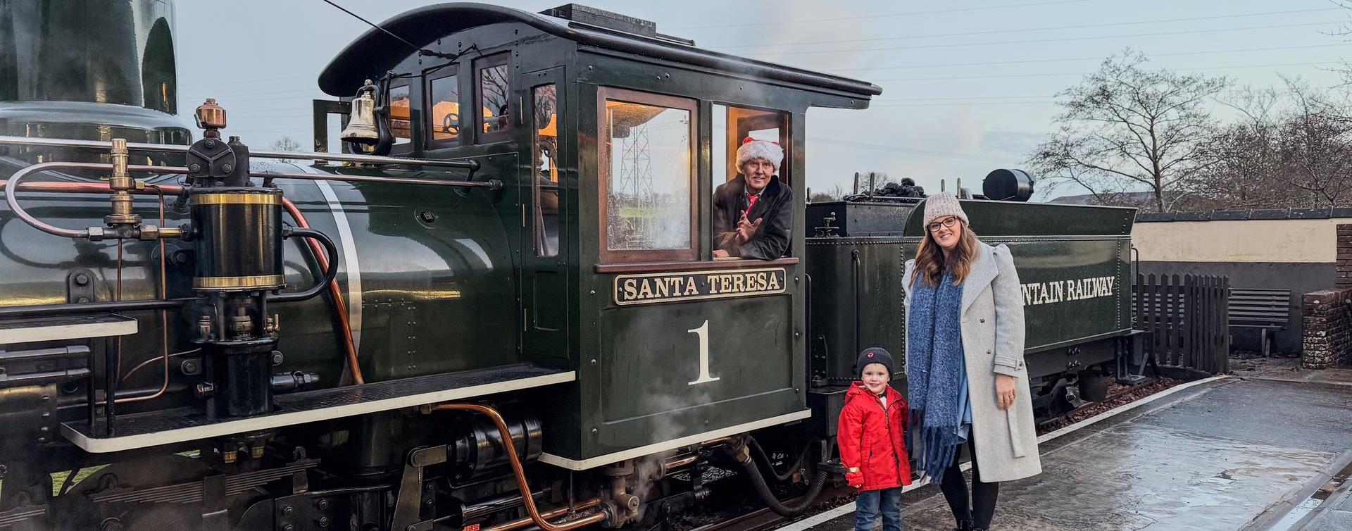 A mother and child beside a steam engine as the driver in a Santa hat waves from the cab