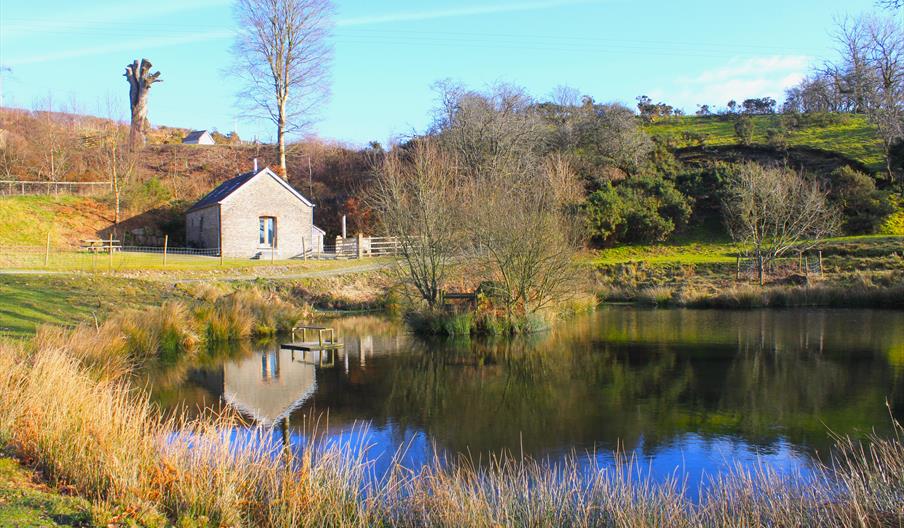 A stone and slate barn sits across from a pond, its reflection clear on water. Trees and grass surround the pond
