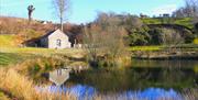 A stone and slate barn sits across from a pond, its reflection clear on water. Trees and grass surround the pond